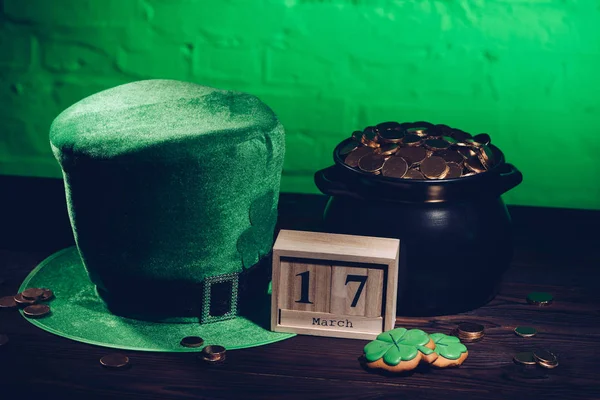 Calendario, galletas en forma de trébol, sombrero irlandés verde y olla con monedas de oro en la mesa de madera - foto de stock