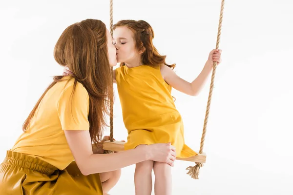 Hija sentada en columpio y besándose madre aislada en blanco - foto de stock