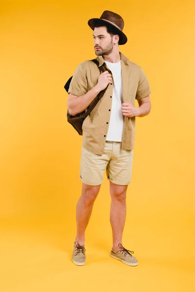 Vue pleine longueur de beau jeune homme en short et chapeau tenant sac à dos et regardant loin isolé sur jaune — Photo de stock