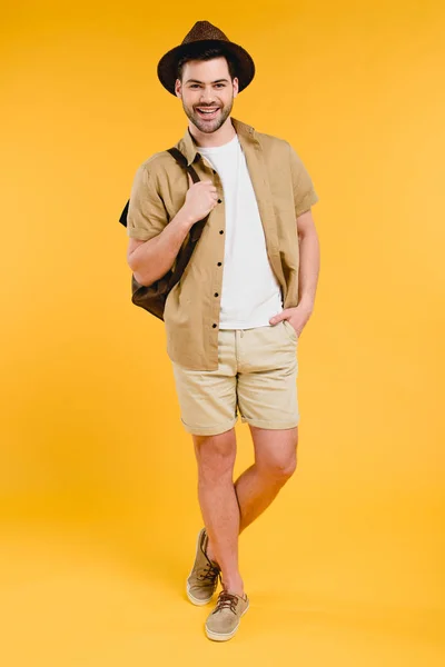 Full length view of handsome young man in shorts and hat holding backpack and smiling at camera isolated on yellow — Stock Photo