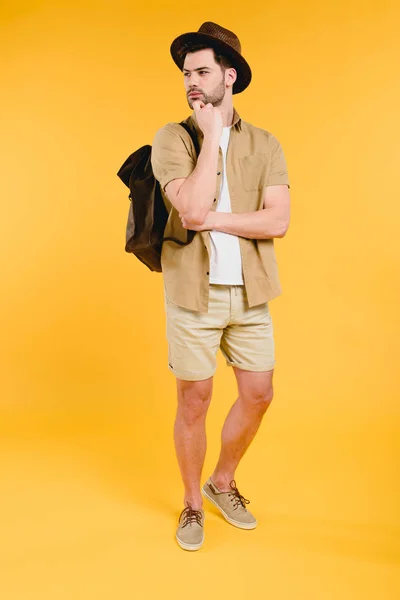 Вид в полный рост молодого человека в шортах с рюкзаком, отводящего взгляд в сторону, изолированный от желтого — стоковое фото