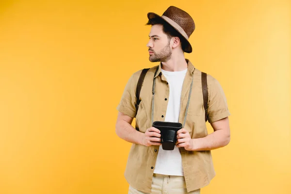Jovem de chapéu com mochila segurando câmera e olhando para longe isolado no amarelo — Fotografia de Stock