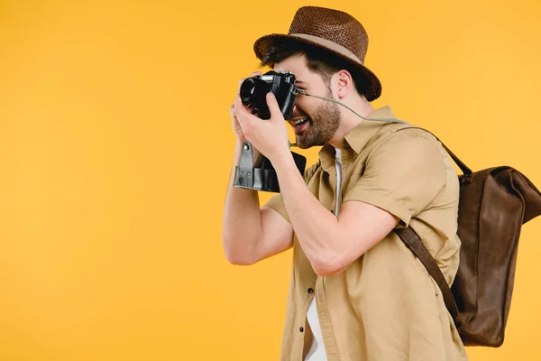 Seitenansicht eines jungen Mannes mit Hut und Rucksack, der mit einer Kamera isoliert auf gelb fotografiert — Stockfoto