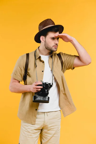 Молодой мужчина путешественник в шляпе держит камеру и смотрит в сторону изолированы на желтый — стоковое фото
