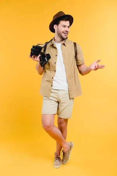 Handsome smiling young man in hat holding camera and looking away isolated on yellow — Stock Photo