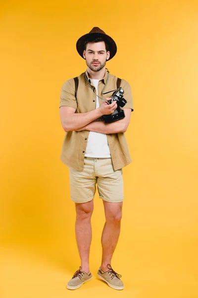 Bonito jovem viajante em shorts e chapéu segurando câmera isolada no amarelo — Fotografia de Stock