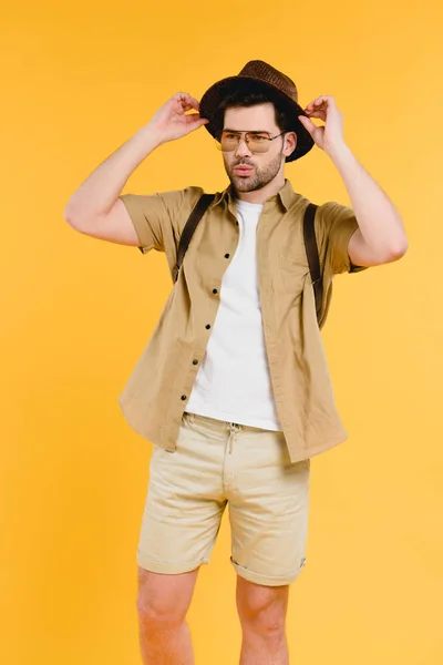 Retrato de joven guapo en gafas de sol y pantalones cortos ajustando sombrero aislado en amarillo - foto de stock