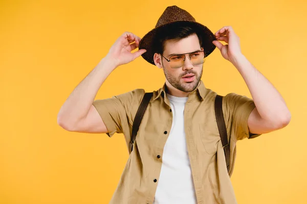 Портрет красивого молодого чоловіка в сонцезахисних окулярах, що регулює капелюх і дивиться ізольовано на жовтий — стокове фото
