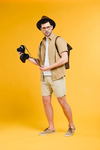 Красивий молодий мандрівник з рюкзаком тримає камеру ізольовано на жовтому — стокове фото