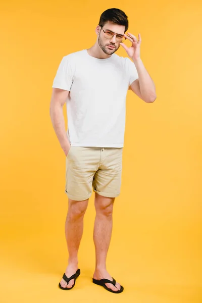 Beau jeune homme en short et tongs ajuster les lunettes de soleil et regarder la caméra isolée sur jaune — Photo de stock