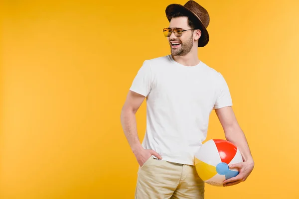 Schöner lächelnder junger Mann mit Hut und Sonnenbrille hält aufblasbaren Ball in der Hand und schaut isoliert auf gelbem Grund weg — Stockfoto