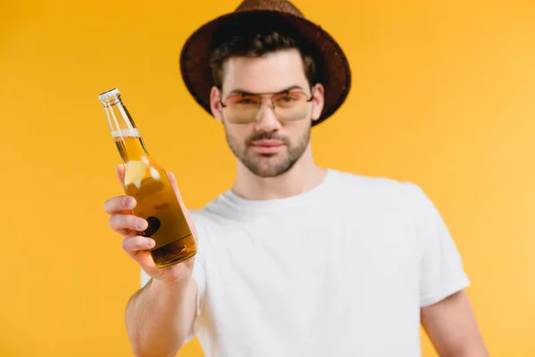 Bello giovane uomo in cappello e occhiali da sole in possesso di bottiglia con bevanda estiva e guardando la fotocamera isolata sul giallo — Foto stock