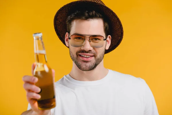 Красивый молодой человек в шляпе и солнцезащитных очках держит бутылку с летним напитком и улыбается в камеру, изолированную на желтый — стоковое фото