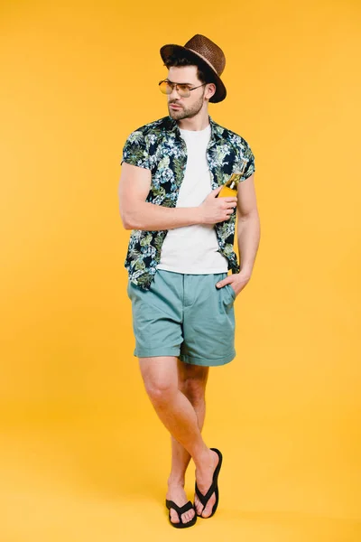 Joven guapo en traje de verano sosteniendo botella de vidrio de bebida refrescante y mirando hacia otro lado aislado en amarillo - foto de stock