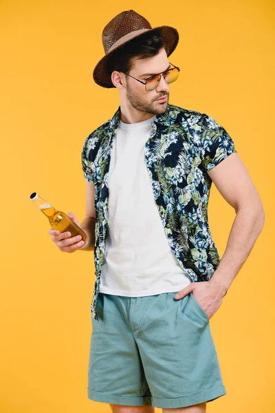Joven con estilo en traje de verano sosteniendo botella de vidrio de bebida de verano y mirando hacia otro lado aislado en amarillo - foto de stock