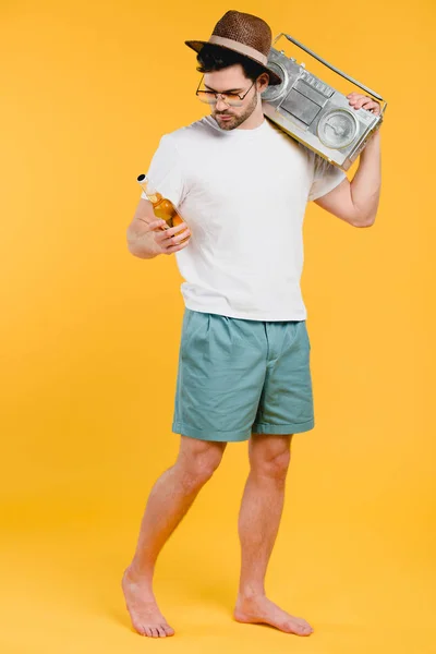 Young barefoot man in shorts holding tape recorder on shoulder and looking at glass bottle of summer drink isolated on yellow — Stock Photo
