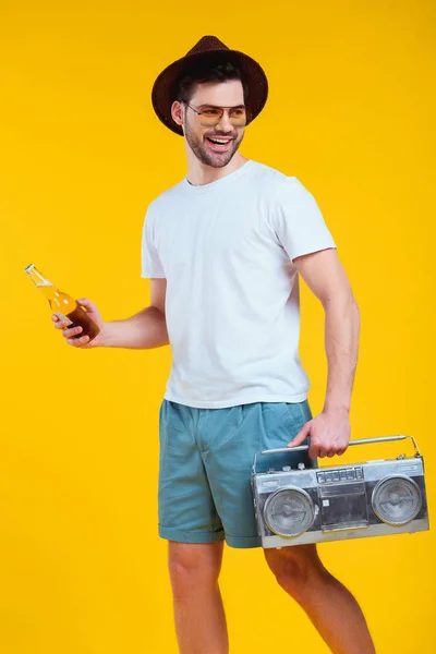 Jeune homme souriant en chapeau et lunettes de soleil tenant magnétophone et bouteille de boisson d'été isolé sur jaune — Photo de stock