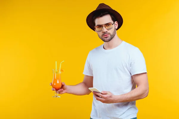 Schöner junger Mann blickt in die Kamera, während er ein Glas Sommercocktail in der Hand hält und das Smartphone isoliert auf gelb benutzt — Stockfoto