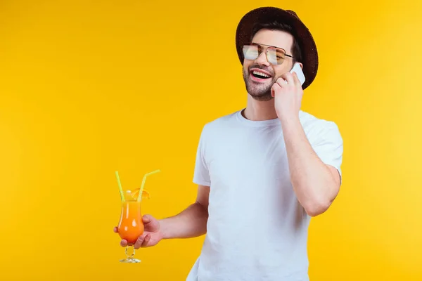 Bel giovane sorridente in cappello e occhiali da sole che regge un bicchiere di cocktail estivo e parla da smartphone isolato su giallo — Foto stock