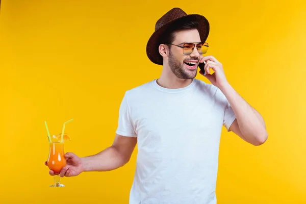 Joven alegre en sombrero y gafas de sol sosteniendo vaso de cóctel de verano y hablando por teléfono inteligente aislado en amarillo - foto de stock