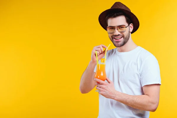 Guapo joven en sombrero y gafas de sol bebiendo cóctel de verano y sonriendo a la cámara aislado en amarillo - foto de stock
