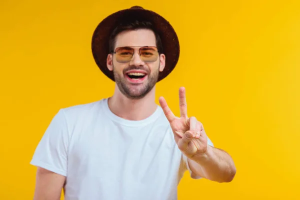 Joven alegre en camiseta blanca, sombrero y gafas de sol mostrando signo de victoria y sonriendo a la cámara aislado en amarillo - foto de stock