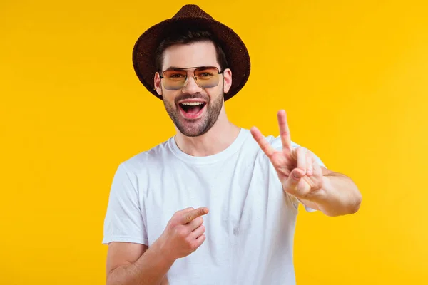 Веселый молодой человек в белой футболке, шляпе и солнцезащитных очках, показывающий знак победы и указывающий пальцем изолированным на желтый — стоковое фото