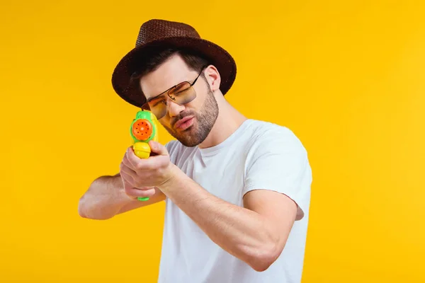 Jeune homme barbu en chapeau et lunettes de soleil tir avec pistolet à eau isolé sur jaune — Photo de stock