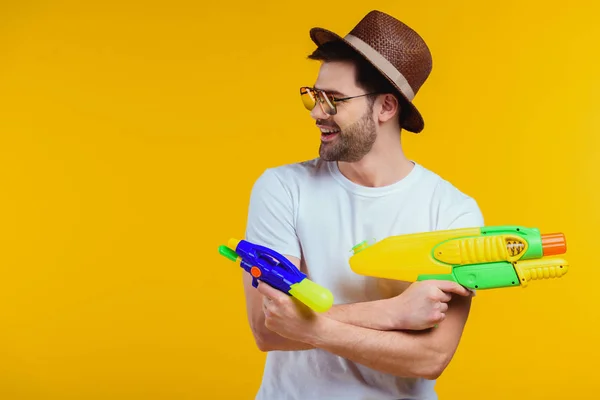 Sonriente joven con sombrero y gafas de sol jugando con pistolas de agua aisladas en amarillo - foto de stock