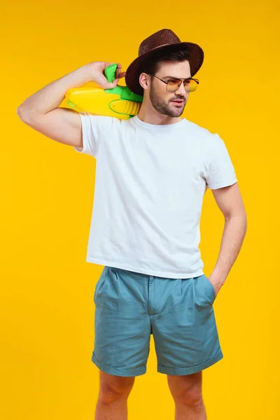 Bel giovane barbuto in abito estivo con pistola ad acqua e guardando lontano isolato sul giallo — Foto stock