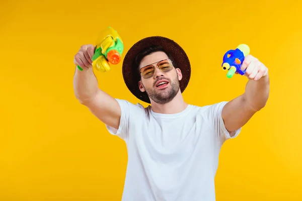 Élégant jeune homme en chapeau et lunettes de soleil tenant des pistolets à eau et regardant la caméra isolée sur jaune — Photo de stock