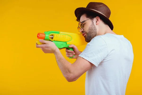 Seitenansicht eines bärtigen jungen Mannes mit Hut und Sonnenbrille, der mit einer Wasserpistole auf Gelb schießt — Stockfoto