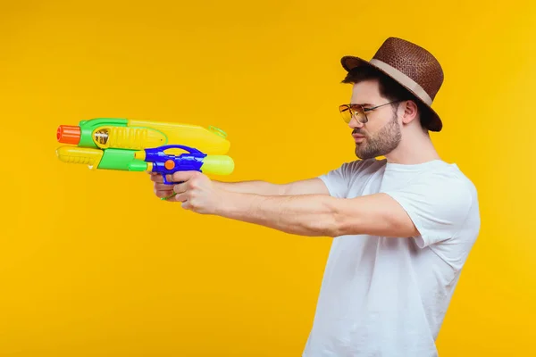 Vue latérale du jeune homme tenant un pistolet à eau et regardant loin isolé sur jaune — Photo de stock