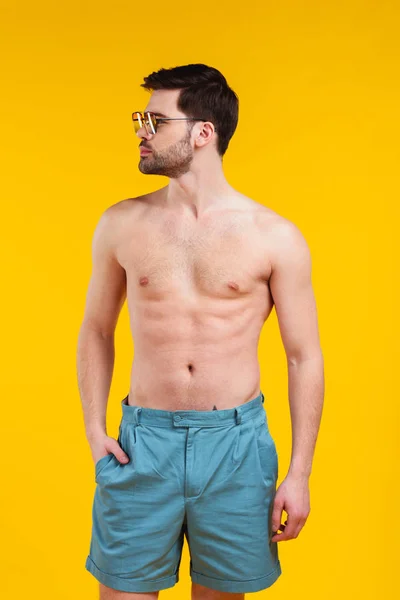 Schöner, hemdloser junger Mann in Shorts und Sonnenbrille, der mit der Hand in der Tasche steht und isoliert auf gelbem Grund wegschaut — Stockfoto