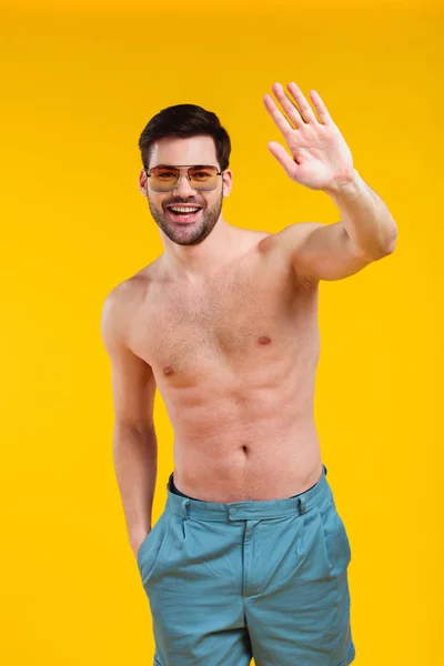 Bel ragazzo senza maglietta in pantaloncini e occhiali da sole agitando la mano e sorridendo alla fotocamera isolata sul giallo — Foto stock