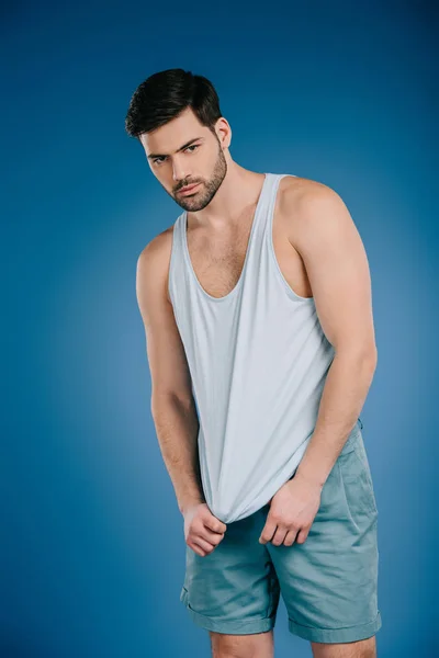 Handsome bearded young man in shorts and white singlet looking at camera on blue — Stock Photo