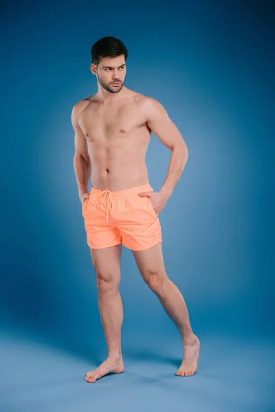 Visão comprimento total do homem descalço bonito em shorts de pé com as mãos nos bolsos e olhando para longe em azul — Fotografia de Stock