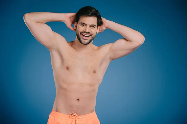 Joven sin camisa guapo de pie con las manos detrás de la cabeza y sonriendo a la cámara en azul - foto de stock