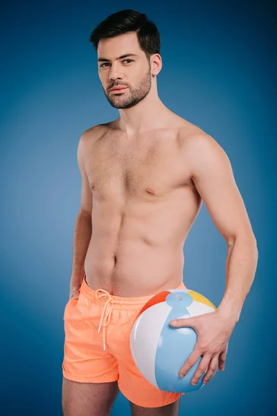 Handsome young shirtless man in shorts holding inflatable ball and looking at camera on blue — Stock Photo