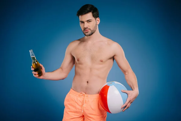 Guapo joven en pantalones cortos sosteniendo botella de bebida de verano y bola inflable y mirando a la cámara en azul - foto de stock
