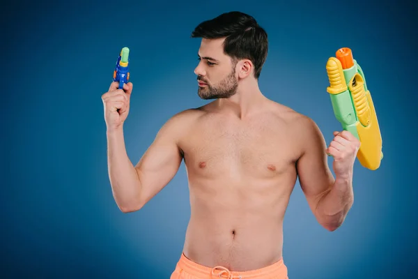 Barbudo joven desnudo pecho hombre sosteniendo armas de agua en azul - foto de stock