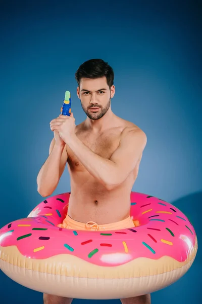 Hombre joven y guapo sin camisa en anillo inflable sosteniendo pistola de agua y mirando a la cámara en azul - foto de stock