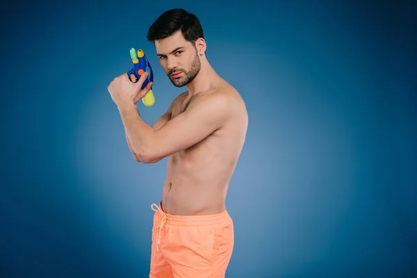 Schöner junger Mann in kurzen Hosen mit Wasserpistole und Blick in die Kamera auf blau — Stockfoto