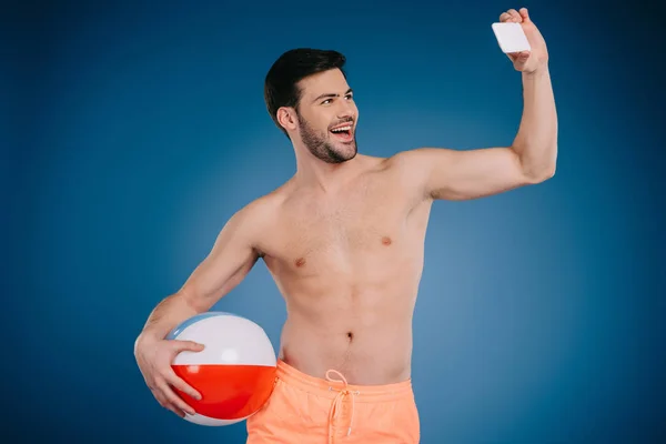 Giovane uomo sorridente in pantaloncini che tiene palla da spiaggia e si fa selfie con smartphone su blu — Foto stock