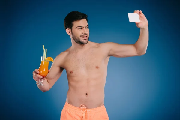Apuesto joven sin camisa en pantalones cortos sosteniendo vaso de cóctel de verano y tomar selfie con teléfono inteligente en azul - foto de stock