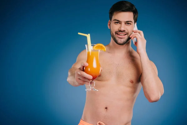 Guapo sin camisa joven sosteniendo vaso de cóctel de verano y hablando por teléfono inteligente en azul - foto de stock