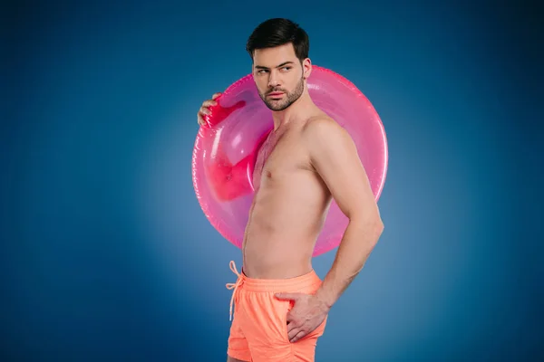 Joven guapo en pantalones cortos sosteniendo anillo de natación y mirando hacia otro lado en azul - foto de stock