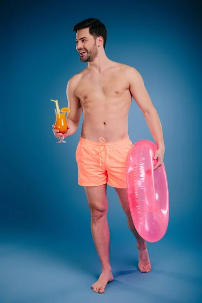 Joven sonriente en pantalones cortos sosteniendo vaso de cóctel de verano y anillo inflable en azul - foto de stock