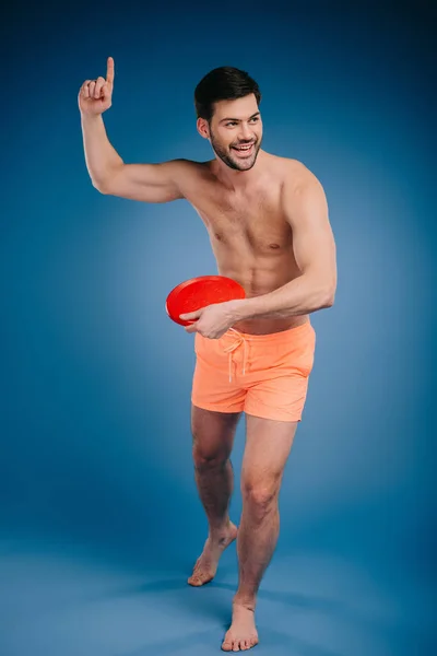 Full length view of smiling young barefoot man in shorts holding flying disk and looking away on blue — Stock Photo