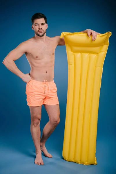 Joven guapo en pantalones cortos de pie con colchón de natación y mirando a la cámara en azul - foto de stock